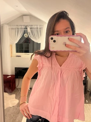 Ljusrosa blus med volangärmar - Supersöt ljusrosa blus med v-ringning och volangdetaljer vid ärmarna. Blusen är luftig och har fina rynkade partier upptill samt spetsliknande detaljer längs axlarna. Perfekt för en fräsch och feminin look.