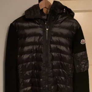 Svart Moncler cardigan - Snygg svart pufferjacka från Moncler med quiltad front och huva. Ärmarna är stickade och har en dragkedjeficka på vänster arm med Moncler-logga. Jackan har två fickor fram och stängs med dragkedja. Perfekt för kyliga dagar.