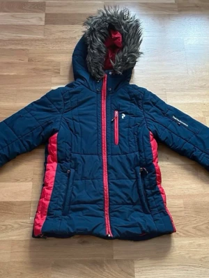 Peak Performance vinterjacka - Mörkblå vinterjacka från Peak Performance med röda detaljer och dragkedjor. Jackan har huva med fuskpäls och flera fickor. Perfekt för kalla dagar och har en sportig vibe. Snygg passform och tydliga logodetaljer. Sprucken vänster ficka (se bild).