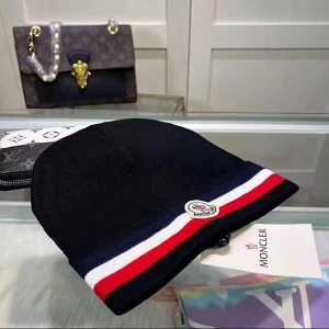 Moncler svart mössa med ränder - Svart Moncler-mössa i stickat material med klassiska ränder i blått, vitt och rött längs kanten. Mössan har Moncler-logga framtill och är perfekt för dig som vill ha en stilren och sportig accessoar till vintern.