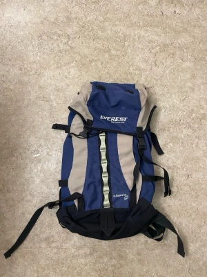 Blå och beige Everest ryggsäck - Säljer en stor och praktisk ryggsäck från Everest med blå och beige paneler, svarta detaljer och flera justerbara remmar. Perfekt för dig som gillar äventyr och behöver mycket packutrymme. Materialet är slitstark polyester och designen är klassisk med lock och spännen.