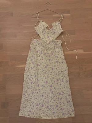 Blommig långklänning med snörning - Säljer en ljusgul långklänning med lila och rosa blommigt mönster. Klänningen har smala axelband, snörning i ryggen och omlott-detaljer vid bysten. Perfekt för sommarens alla tillfällen, superfin och romantisk design.