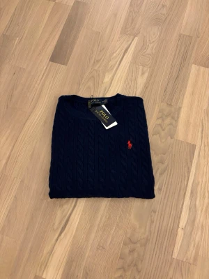 Mörkblå stickad tröja Polo Ralph Lauren - Snygg mörkblå stickad tröja från Polo Ralph Lauren i klassisk kabelstickad design. Tröjan har rund hals, långa ärmar och en röd broderad logga på bröstet. Perfekt för dig som gillar stilrena och tidlösa plagg med premiumkänsla.