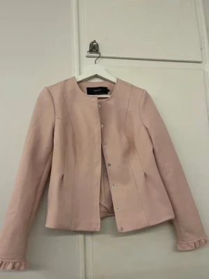 Ljusrosa jacka - Säljer en ljusrosa kavaj från Vero Moda med rund halsringning och tryckknappar framtill. Jackan har långärmade ärmar med gulliga volangdetaljer vid ärmsluten. Materialet är mjukt och har en lätt struktur som ger en stilren look.