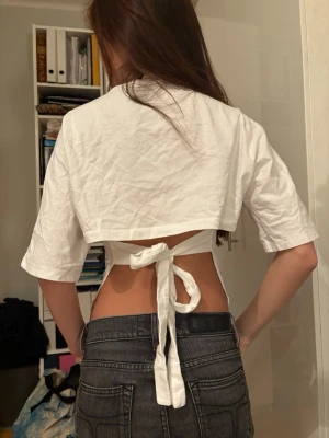Nelly topp - Säljer en vit croppad topp med korta ärmar och snygg knytning i ryggen. Toppen har en enkel och stilren design med rund hals och är tillverkad i ett lätt bomullsmaterial. Perfekt för dig som gillar trendiga och unika detaljer.
