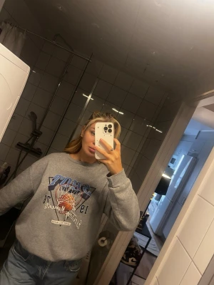 Grå sweatshirt med baskettryck - Säljer en grå sweatshirt med coolt baskettryck framtill i blått, vitt och orange. Tröjan har rund hals, långa ärmar och en avslappnad passform. Perfekt för dig som gillar sportiga vibes och streetstyle.