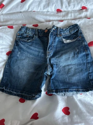 Blå jeansshorts från 157 Sunny Star - Säljer ett par klassiska blå jeansshorts från 157 Sunny Star. De har normal passform, fem fickor och knapp- samt dragkedja framtill. Shortsen är tillverkade i bomull och har en snygg tvättad look med slitningar. Perfekta för chill dagar när du vill vara bekväm och ändå se fräsch ut.