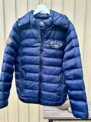 Mörkblå pufferjacka Polo Club S - Snygg mörkblå pufferjacka från Polo Club med broderad logga på bröstet och ärmen. Jackan har huva med tryck, två fickor med dragkedja och quiltad design. Perfekt för kalla dagar.