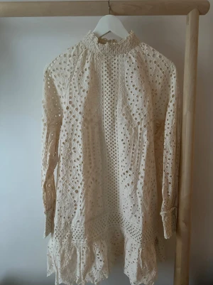 Vit klänning från H&M - Supersöt vit kortklänning från H&M i storlek XS. Klänningen har långärmade ärmar, hög hals och är täckt av broderad spets med hålmönster och volangdetaljer längst ner.