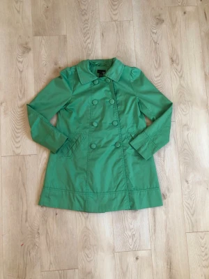 Grön trenchcoat - Snygg grön trenchcoat från H&M med dubbelknäppning och klassisk krage. Jackan har två fickor fram och en stilren slits bak. Materialet är slätt och känns lätt, perfekt för vår och höst. Färgen är klar grön och modellen är något vid.