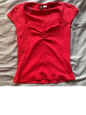  Supersöt Röd ribbad topp med rynk framtill - Säljer en röd ribbad topp från H&M i mjuk bomull. Toppen har korta ärmar, v-ringad hals med virkad kant och snygg rynkdetalj framtill. Perfekt till jeans eller kjol för en enkel men trendig look.