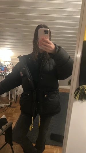 Svart pufferjacka från Fjällräven - Säljer en svart pufferjacka från Fjällräven med gul snörning och guldiga knappar. Jackan har en hög krage, två stora fickor framtill och ett broderat Fjällräven-märke på bröstet. Perfekt för kalla dagar och riktigt snygg till streetwear. Nypris 10 000 