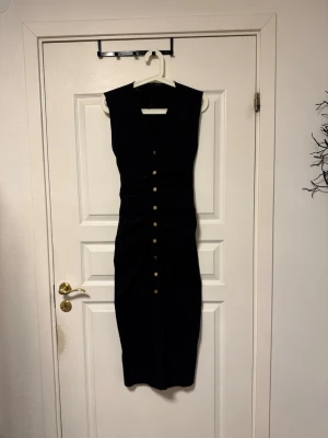 Svart långklänning med knappar - Snygg svart långklänning med v-ringad hals och ärmlös design. Klänningen har guldiga knappar framtill och en figurnära passform med rynkade detaljer vid midjan. Perfekt för dig som gillar stilrena och eleganta plagg.