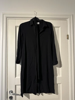 Svart skjortklänning från VILA - Svart skjortklänning från VILA med klassisk krage och knappar hela vägen framtill. Klänningen har trekvartsärm och en rak, lite loose passform. Finns ett svart skärp/band som är avtagbar.
