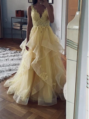 Ljusgul långklänning med spetsdetaljer - Säljer en drömmig ljusgul långklänning med v-ringad spetsliv och volangkjol i lager på lager. Klänningen har ärmlös design och vackra broderade blommor på både liv och kjol. Perfekt för bal eller student.