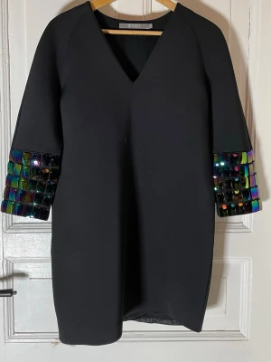 Svart klänning med paljettdetaljer Tiger of Sweden  - Svart kortklänning från Tiger of Sweden med v-ringad hals och trekvartsärm. Ärmarna har stora, glittriga paljetter i regnbågsfärger som ger en cool och unik look. Klänningen har en rak passform och är tillverkad i ett slätt syntetmaterial. Inga synliga fel. Djur finns i hemmet. Mått: midja: 45 cm längd: 82 cm 