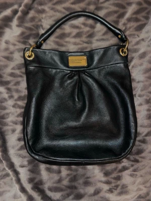 Svart axelväska Marc by Marc Jacobs - Säljer en svart axelväska från Marc by Marc Jacobs, Classic Q Hiller i mjukt skinn med guldfärgade metalldetaljer och en snygg logoplatta framtill. Väskan har både kort och lång rem som man får med. På de två sista bilderna visas en likadan väska som säljs på Sellpy för 940 kr men i sämre skick. Väskan är köpt här på Plick. Skriv för fler bilder eller om du funderar över något❤️