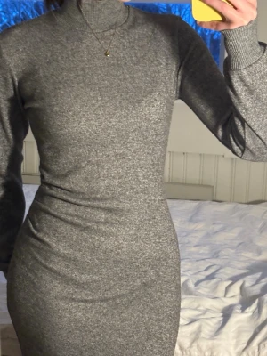 Grå stickad långärmad klänning - Säljer en grå, stickad klänning med polokrage och lång ärm. Klänningen har en kroppsnära passform och är i mjukt material som passar perfekt till kyligare dagar. Enkel och stilren design utan mönster eller extra detaljer.
