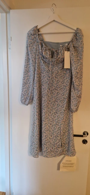 Blommig ljusblå klänning från Adoore - Supersöt ljusblå klänning från Adoore med blommigt mönster i vitt och orange. Klänningen har lång ärm, snörning vid bysten och en liten slits framtill. Materialet känns lätt och luftigt, perfekt för varmare dagar.