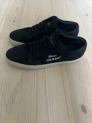 Svarta Saint Laurent sneakers - Säljer ett par svarta Saint Laurent sneakers med vit sula och handskriven logga på sidan. Skorna har snörning, rund tå och är tillverkade i canvas och skinn. Klassisk och stilren modell med diskreta detaljer och guldtext på plösen. Sulan har släppt lite men inget märkvärdigt 
