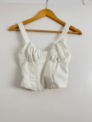 Vit korsett-topp från Bershka - Snygg vit korsett-topp från Bershka med breda axelband och rynkade kupor. Toppen har smockad rygg för extra komfort och en croppad passform. Perfekt att styla med jeans eller kjol för en trendig look.