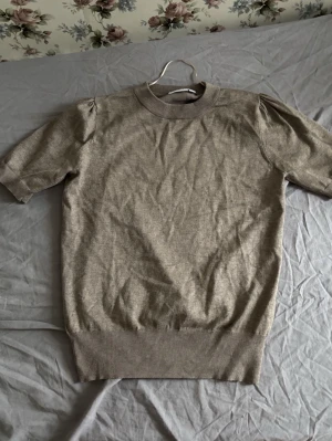 Beige stickad topp med puffärm  - Säljer en beige stickad topp med korta puffärmar och rund hals från Saint Tropez. Toppen har ribbade muddar vid hals och nederkant, vilket ger en snygg och bekväm passform. Perfekt att matcha med jeans eller kjol för en enkel och stilren look.