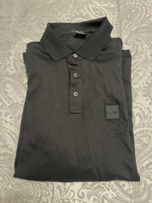 Svart långärmad pikétröja från BOSS - Svart långärmad pikétröja från BOSS i storlek M. Tröjan har klassisk krage, tre knappar framtill och en snygg BOSS-logga på bröstet. Materialet är mjukt och känns skönt mot huden, perfekt för dig som gillar stilrena och enkla plagg.