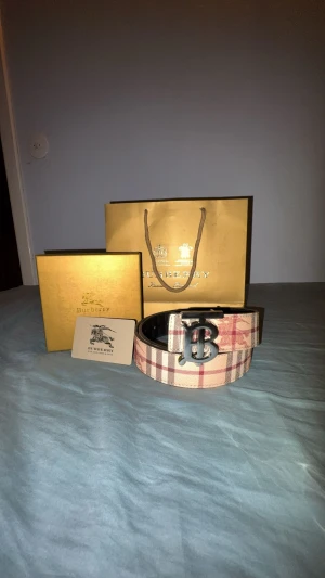 Burberry rutigt skärp med BB-spänne - Säljer ett Burberry bälte i klassiskt beige och rött rutigt mönster med silverfärgat BB-logga som spänne. Bältet är gjort i skinn och har den ikoniska Burberry-rutan med röda och svarta detaljer. Perfekt accessoar för att lyfta din outfit.