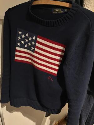 Mörkblå stickad tröja Polo Ralph Lauren - Säljer en mörkblå stickad tröja från Polo Ralph Lauren med stor amerikansk flagga på bröstet och RL-broderi. Tröjan har rund hals, ribbade muddar och lång ärm. Perfekt för dig som gillar klassisk och cool stil.