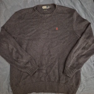 Mörkblå stickad tröja Polo Ralph Lauren - Mörkblå stickad tröja från Polo Ralph Lauren i 100% bomull. Klassisk rund halsringning, ribbade muddar och nederkant. Liten röd broderad logga på bröstet ger en clean och stilren look. Perfekt för lager-på-lager under kyliga dagar.