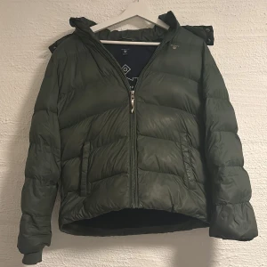 Mörkgrön pufferjacka från GANT - Snygg mörkgrön pufferjacka från GANT med huva och dragkedja framtill. Jackan har en quiltad design och ribbade muddar vid ärmslut. Perfekt för kalla dagar och har en cool, sportig vibe. Passar dig som gillar stilrena och funktionella jackor.