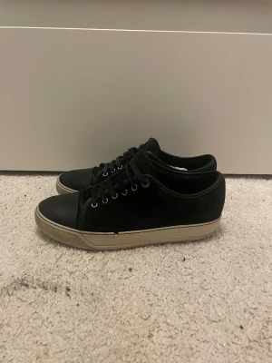 Lanvin - Svarta sneakers med ovandel i mocka och tåparti i skinn. Skorna har svarta snören, silverfärgade öljetter och en beige platt sula. Klassisk och stilren design som passar till det mesta. Perfekta för dig som gillar en enkel men snygg look. ‼️SMÅ DEFEKTER VID CAP TOEN‼️