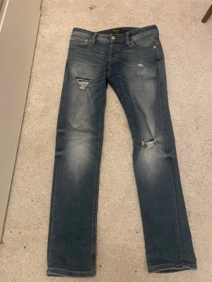 Slim glenn Jack and Jones! - Säljer ett par blå jeans från Jack & Jones med snygga slitningar och hål på låret och knät. Jeansen har klassisk femficksdesign, gylf med dragkedja och knapp, samt en skinnpatch bak i midjan. Passformen är rak och materialet är mjukt denim.