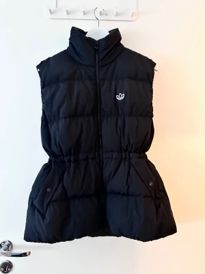 Svart pufferväst från Adidas - Svart pufferväst från Adidas med hög krage och dragkedja framtill. Västen har ett broderat Adidas-logo på bröstet och två fickor med knappstängning. Perfekt för lager-på-lager och chill stil.