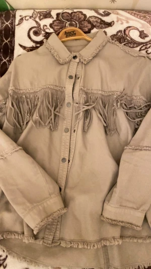 Beige overshirt med fransar från Zara - Snygg beige overshirt från Zara med coola fransdetaljer och silvriga nitar längs bröstet. Overshirten har knappar framtill, råa kanter och lång ärm. Materialet är kraftig bomull som ger en schysst struktur. Perfekt för dig som gillar western vibes och statement-plagg.