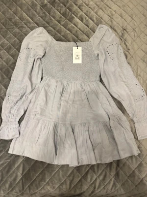 Ljusgrå volangklänning DW Air S - Supersöt ljusgrå kortklänning från DW Air med smockad överdel, volangkjol och långa ärmar med broderade detaljer. Klänningen har fyrkantig ringning och är tillverkad i ett mjukt, luftigt material som känns perfekt för vår och sommar.