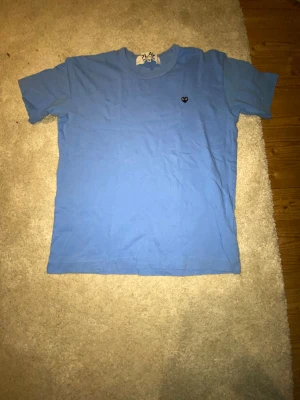Blå Comme des Garçons Play t-shirt M - Blå t-shirt från Comme des Garçons Play i storlek M. Klassisk modell med korta ärmar och rund hals. På bröstet sitter det ikoniska svarta hjärtat med ögon. Tillverkad i mjuk bomull för en skön känsla.