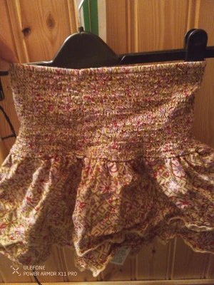 Blommig kortkjol med volang - Supersöt kortkjol med blommigt mönster i beige, rött och rosa. Kjolen har en smockad midja som sitter skönt och volangkant nedtill för extra flärd. Perfekt för dig som gillar boho-vibe och färgglada detaljer. 