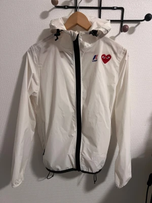 Vit vindjacka K-Way x Comme des Garçons - Selling authentic comne des garcobs jacket. Snygg vit vindjacka från K-Way i samarbete med Comme des Garçons PLAY. Jackan har huva, svart dragkedja och snörning nertill. På bröstet finns det klassiska röda hjärtat med ögon och K-Way-loggan. Perfekt för dig som gillar streetwear och coola detaljer.