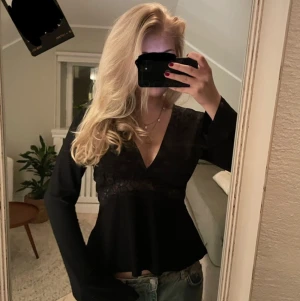 Svart blus med spets och v-ringning - Säljer en svart blus med lång ärm och v-ringad hals. Toppen har snygga spetsdetaljer över midjan. Modellen är något utsvängd nedtill och har en normal passform. Perfekt för dig som gillar stilrena och trendiga plagg. Aldrig använd 