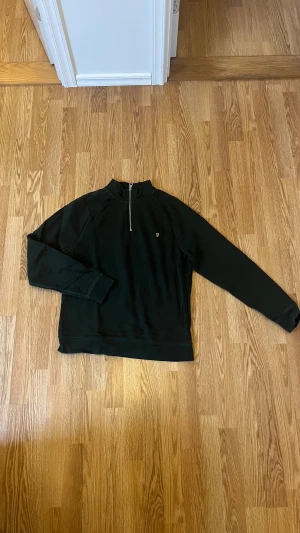 Grön half zip tröja från Farah - Svart långärmad tröja från Farah i 100% ekologisk bomull. Tröjan har en snygg half zip-dragkedja framtill och ett litet gult Farah-logo broderat på bröstet. Perfekt för chill dagar och enkel att matcha med jeans eller joggers.