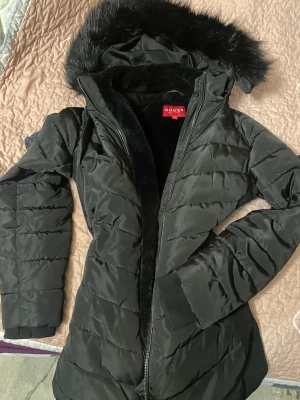 GUESS svart lång pufferjacka  - Svart lång pufferjacka från GUESS med huva och fluffig fuskpäls. Jackan har dragkedja framtill, ribbade muddar och ett snyggt Guess-band i midjan på ryggen. Quiltad design och mjukt foder gör den perfekt för kalla dagar. Snygg och stilren look :)