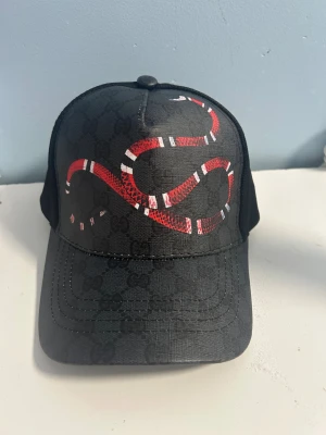 Svart Gucci keps med röd orm - Snygg svart keps från Gucci med ikoniskt GG-mönster och en stor röd och vit orm på framsidan. Kepsen har böjd skärm och mesh på sidorna för extra ventilation. Märket syns tydligt på insidan och etiketten visar att den är tillverkad i Italien.