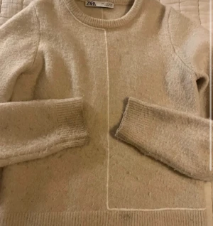 Beige stickad tröja från Zara - Mysig beige stickad tröja från Zara med rund hals och ribbade muddar vid ärmslut och nederkant. Tröjan har en diskret söm framtill som ger en cool detalj. Perfekt för att hålla sig varm under kyliga dagar.