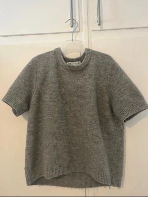 Grå stickad tröja från Zara - Mysig grå stickad tröja från Zara med korta ärmar och rund hals. Tröjan har en enkel, loose passform och ribbade detaljer vid hals, ärmslut och nederkant. Perfekt för lager-på-lager under höst och vinter.