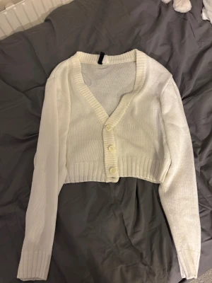 Vit croppad kofta från H&M Divided - Säljer en vit croppad kofta från H&M Divided i storlek S. Koftan har ribbade muddar, v-ringning och tre knappar framtill. Den är stickad och har långa ärmar, perfekt att slänga över en topp för en chill look. Lite nopprig men annars i bra skick💕