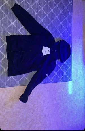 Svart vindjacka från Moncler med huva - Svart vindjacka från Moncler med klassisk logga på ärmen och huva med röd, vit och blå rand. Jackan har dragkedja framtill och är tillverkad i lätt polyester. Perfekt för dig som vill ha en stilren och sportig look.