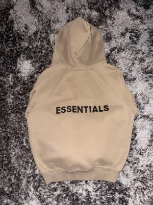 Beige Essentials Fear of God hoodie - Snygg beige hoodie från Essentials Fear of God i storlek S. Tröjan har huva, dragkedja framtill och svart Essentials-tryck på ryggen. Tillverkad i mjuk bomullsmix med ribbade muddar och fickor på sidorna. Perfekt för en chill och stilren look.