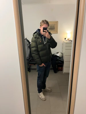 Mörkgrön pufferjacka från Polo Ralph Lauren - Säljer en mörkgrön pufferjacka från Polo Ralph Lauren med huva och orange logga på bröstet. Jackan har flera praktiska fickor med dragkedja. Perfekt för kalla dagar. Använd typ en vinter. Köpt på NK och kvitto finns. Bara att höra av sig om man går någon fundering.