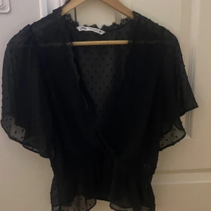 Svart prickig blus från Zara - Säljer en svart blus från Zara perfekt till fester och middagar, i storlek M men är lite mindre i storleken. Blusen är transparent med prickigt mönster, har omlott-design och spetsdetaljer vid halsen. Modellen är luftig med korta ärmar och resår i midjan💕